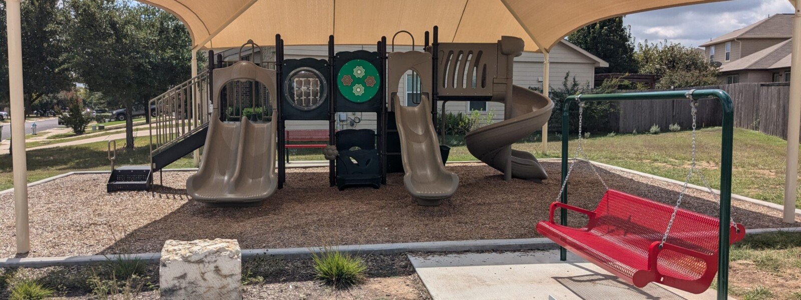 Playground under shade structure, with a multi-person swing in the foreground.